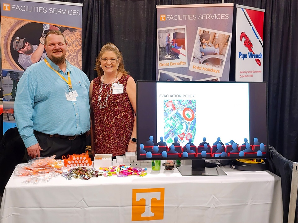 image of two Facilities Services employees smiling behind a table with promotional items at a community job fair