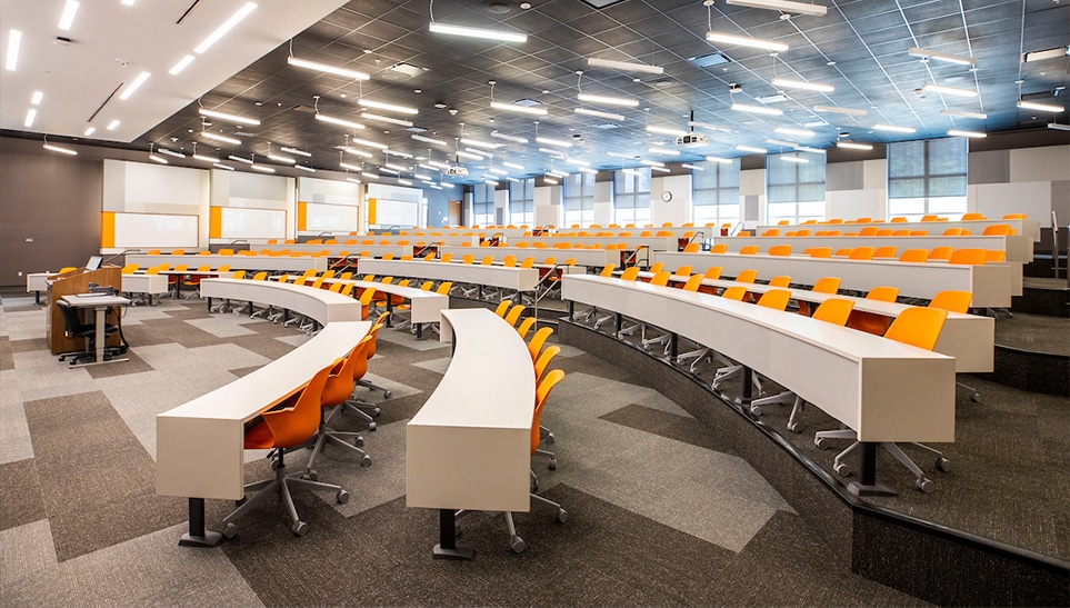photo of a classroom in auditorium style with orange chairs and long desktops all facing the front of the classroom