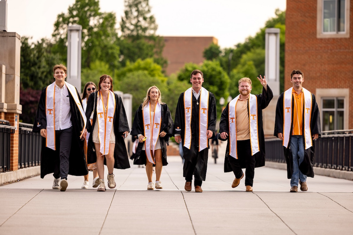 A group of graduates walks across Ped Walkway wearing academic regalia and stoles with the University of Tennessee "T" 