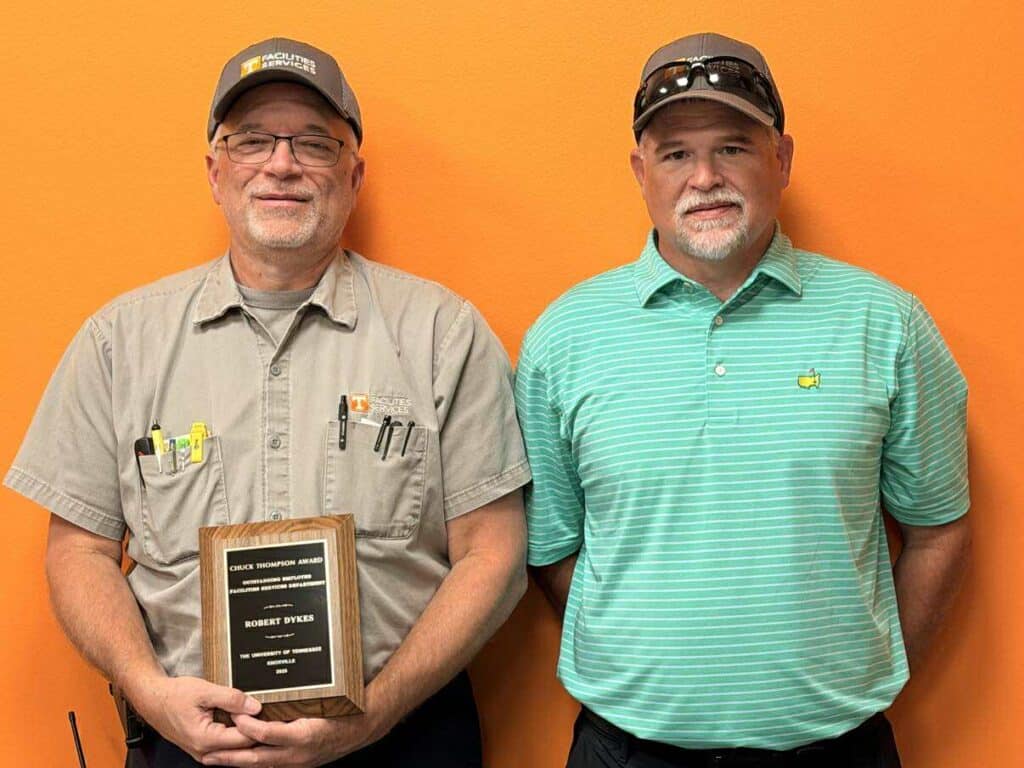 Robert Dykes poses with his plaque and Assistant Vice Chancellor of Facilities Services Operations, Derek Bailey against an orange wall.