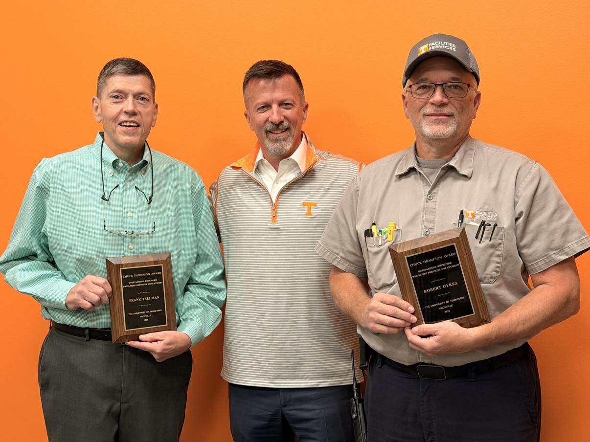 Left to Right: 2025 Chuck Thompson Award winner Frank Tallman, Facilities Services Associate Vice Chancellor Mike Brady, and 2025 Chuck Thompson Award winner Robert Dykes post against an orange wall. The winners are holding their plaques.