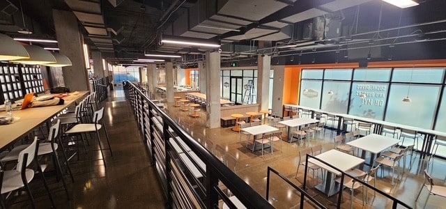 A modern, multi-level common area with industrial design, featuring a long bar, various tables and chairs, concrete pillars, and a large window with orange accents.