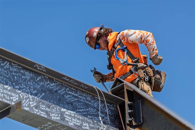 A construction worker in an orange shirt and hard hat works on a steel beam covered in signatures against a clear blue sky.
