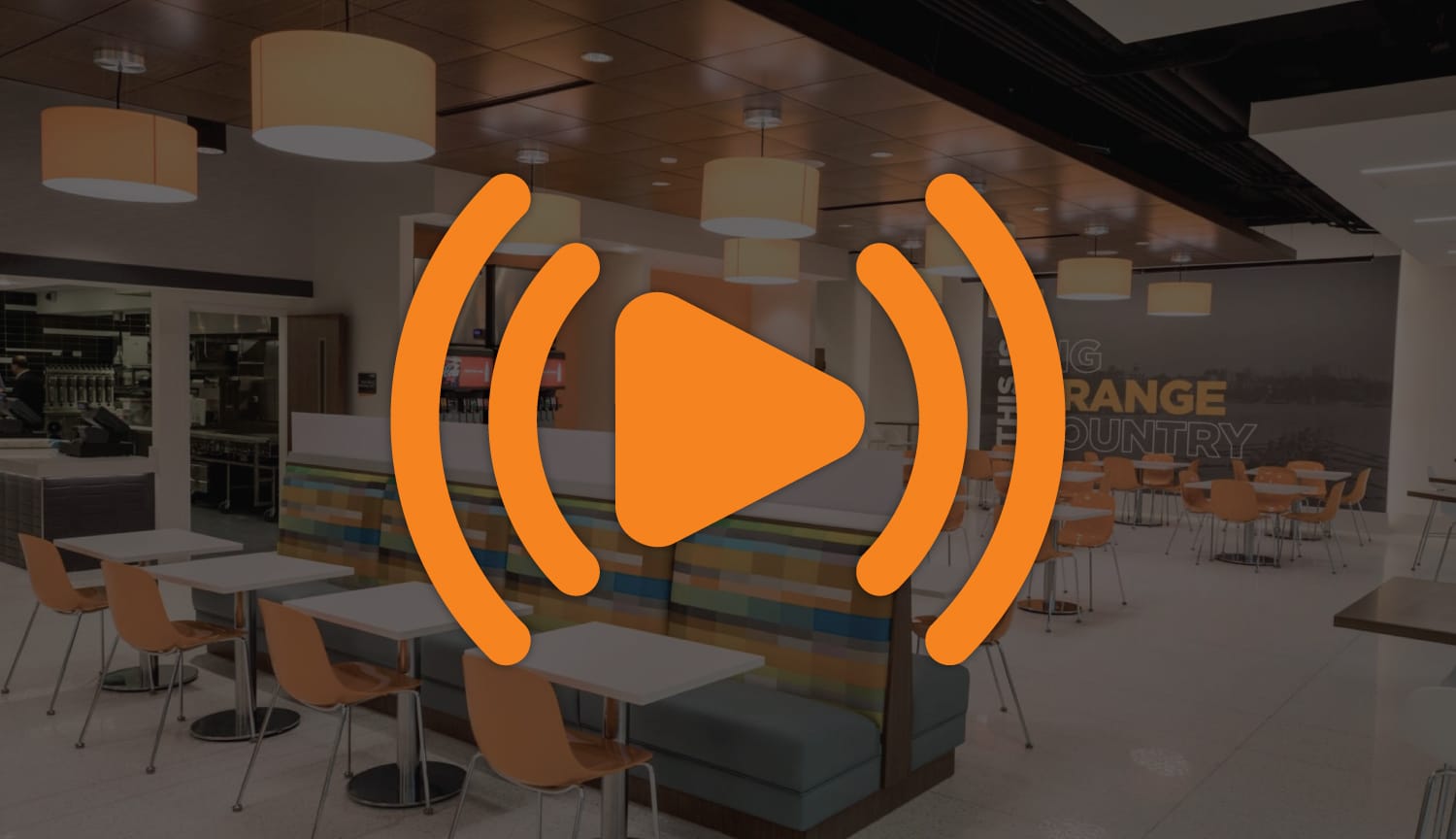 A dark image of a modern cafeteria with orange chairs and colorful booths, overlaid with a large orange play button and audio wave icon.