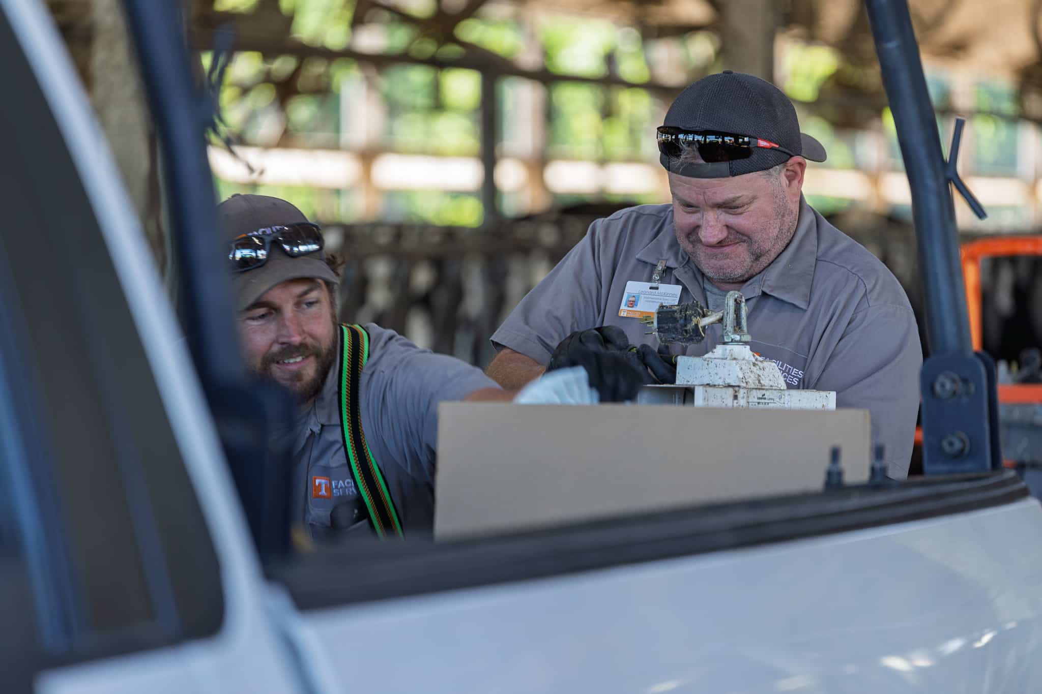 Two men in gray work shirts and caps, one wearing black gloves, are focused on working on a white mechanical part, framed by the interior of a white vehicle.