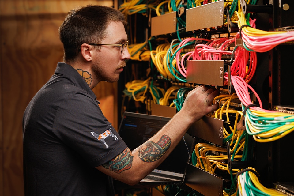 A man with glasses connects cables in a server rack, with a laptop open below.