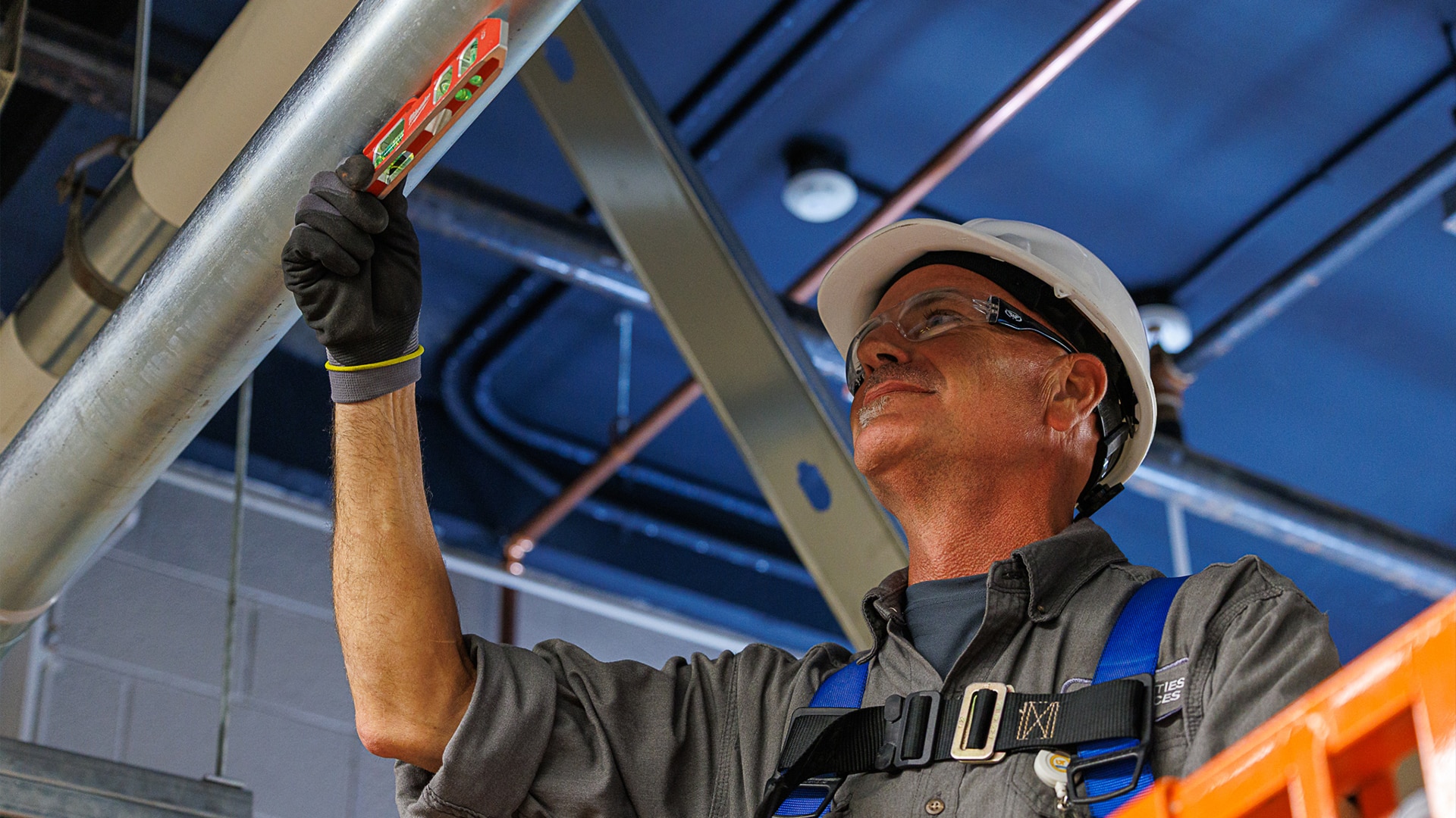 A construction worker in a hard hat and safety glasses uses a magnetic level to check a large metal pipe overhead.