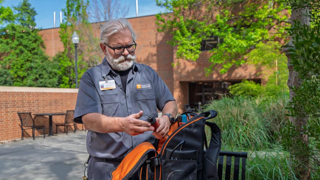 A man with a gray beard and glasses, wearing a gray Facilities Services shirt, holds a tool while standing next to an open orange and black tool backpack outdoors.