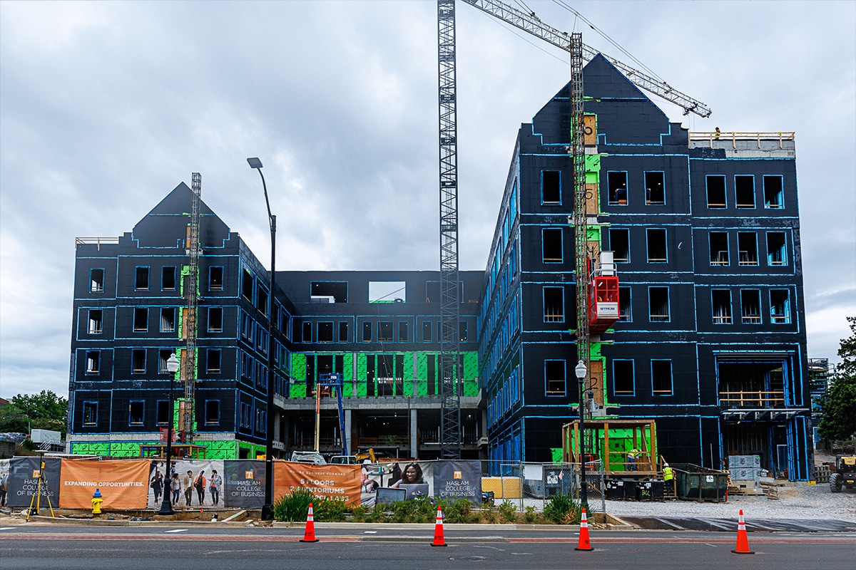 A large building under construction with dark sheathing, a tall tower crane, and a red personnel hoist, viewed from across a street with orange construction fencing and traffic cones.