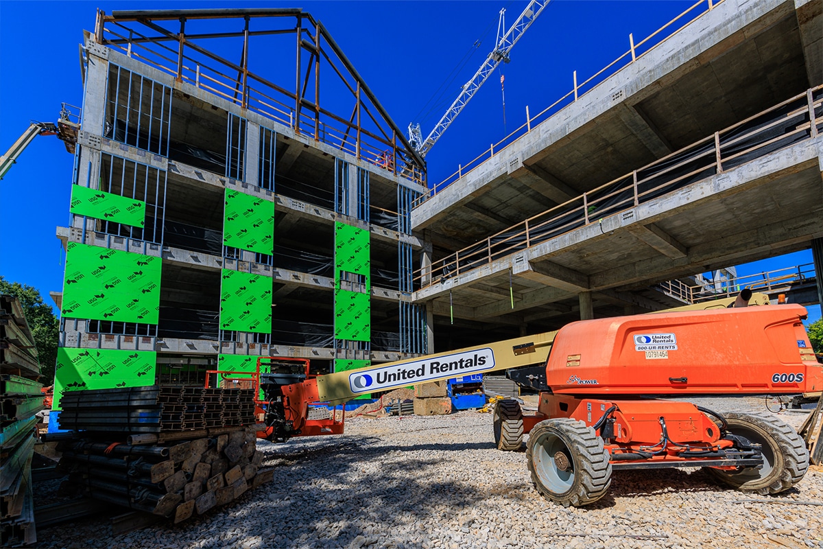 A construction site with two multi-story buildings under construction, a JLG 600S boom lift, and stacks of materials on gravel ground under a blue sky.