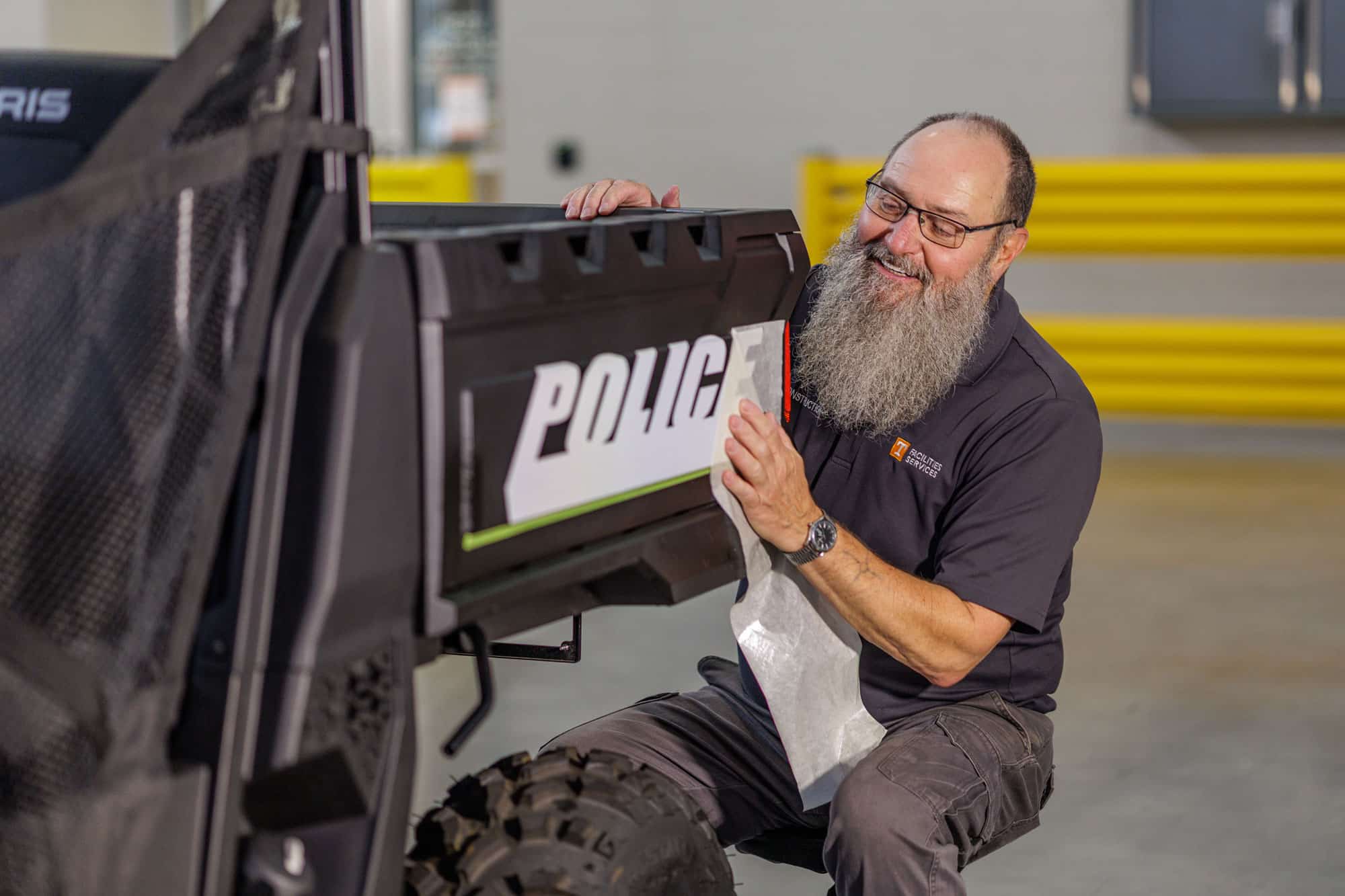 A smiling man with a long grey beard and glasses cleans the 'POLICE' lettering on the side of a black utility vehicle with a white cloth.