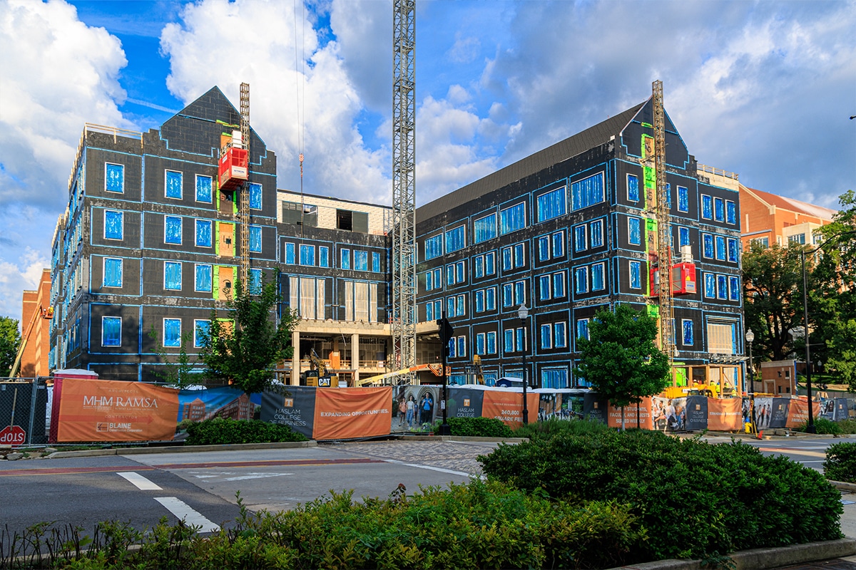 A large building under construction with dark sheathing, a tall tower crane, and a red personnel hoist, viewed from across a street with orange construction fencing and traffic cones.