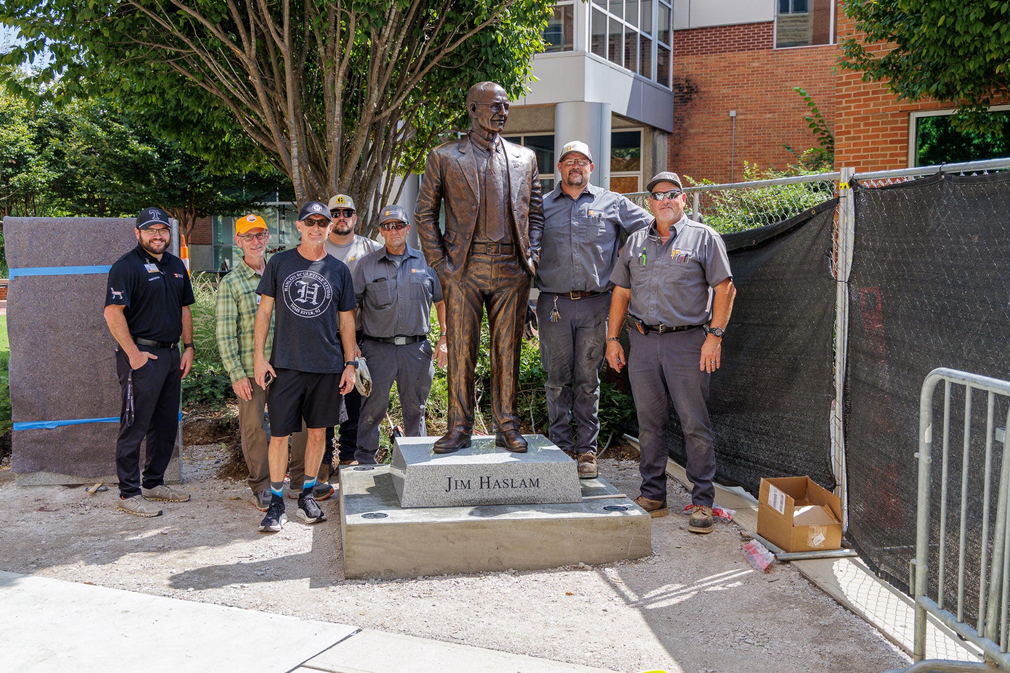 Seven men stand around a bronze statue of Jim Haslam in a suit, which is on a stone base, outdoors on a sunny day.