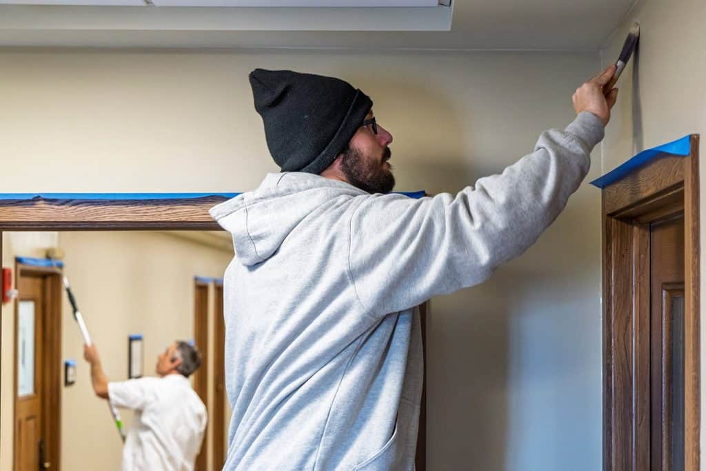man in a hoodie and toboggan uses a paint brush to paint near ceiling of an interior hallway